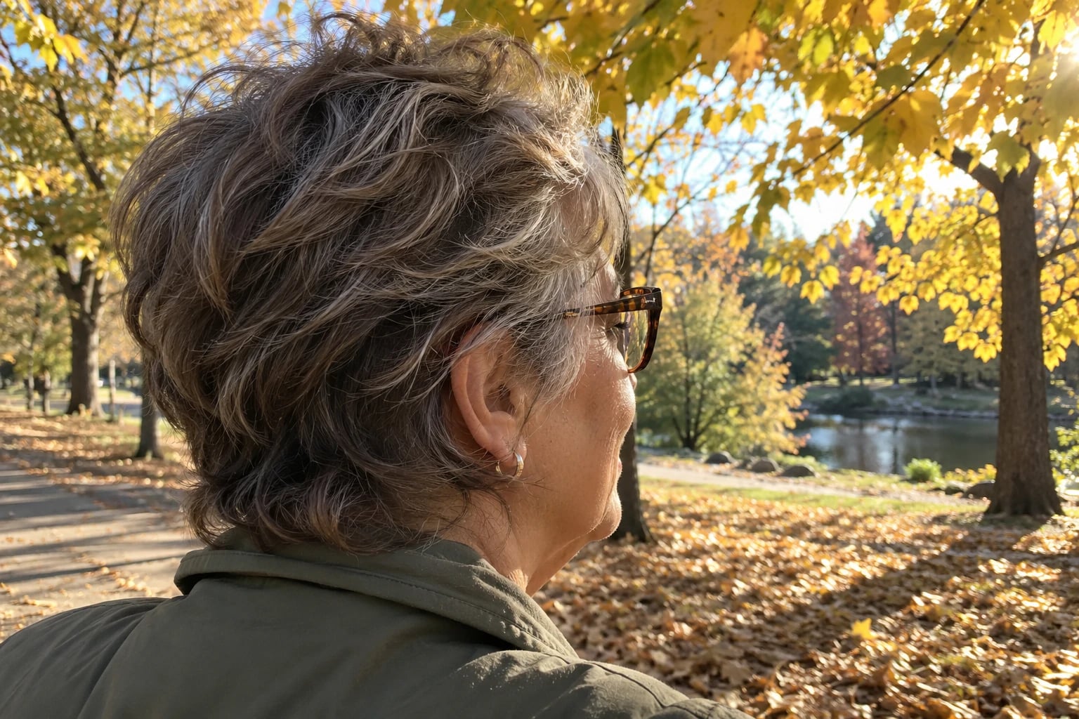 A woman in her sixties with a tousled grey-brown pixie and tortoiseshell glasses gazes across a golden autumn park.