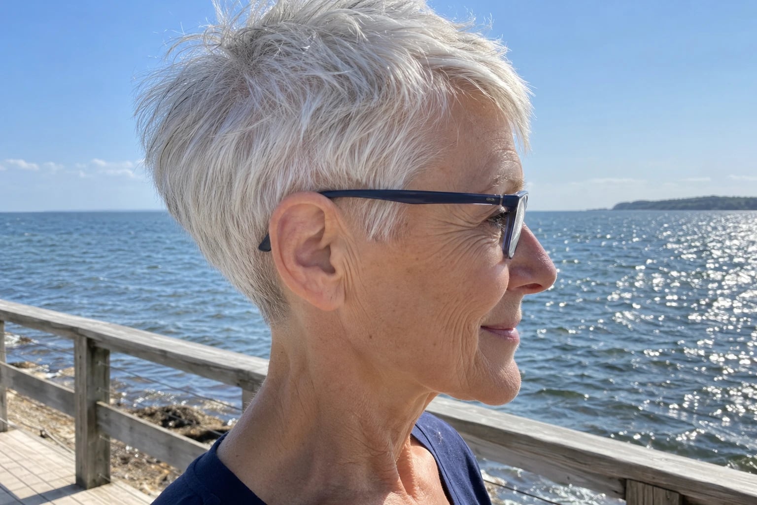 A woman in her sixties with a white pixie cut wears navy rectangular glasses beside the sea.