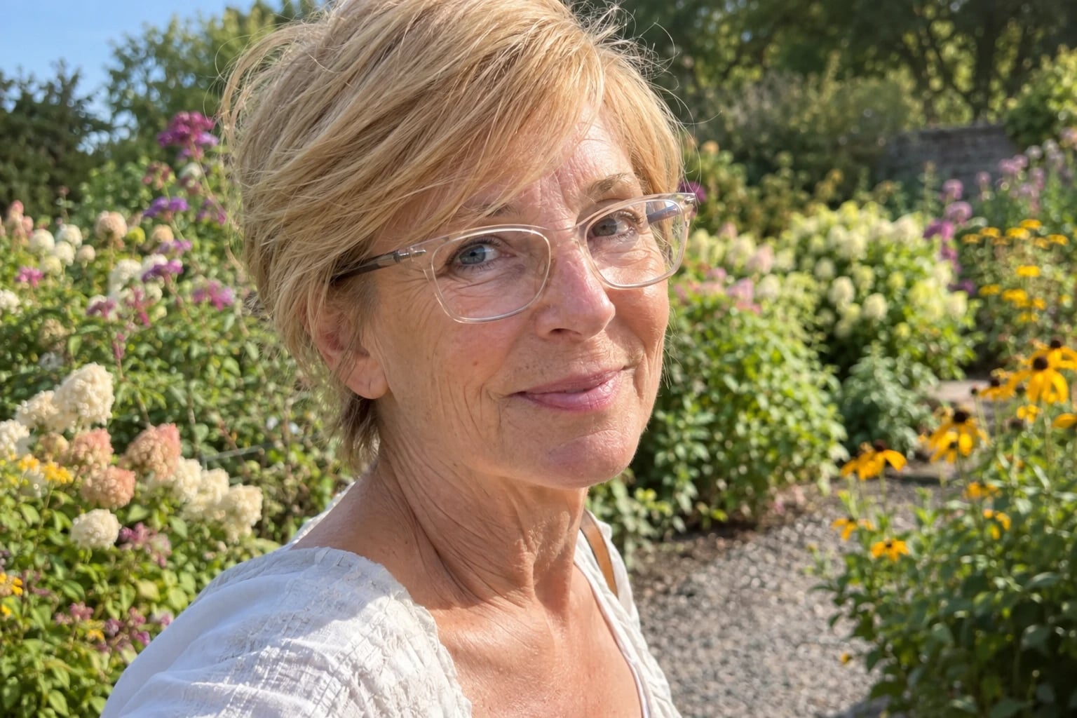 A woman in her sixties wears a honey-blonde side-swept pixie and clear rectangular glasses beside a flower garden path.