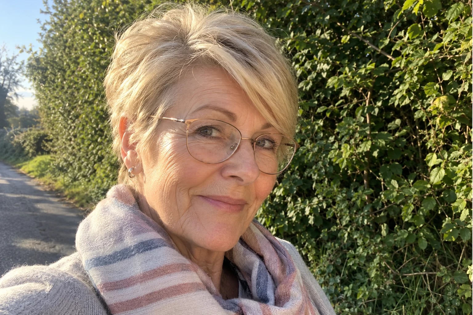 A woman in her sixties with a golden blonde pixie cut and rose-gold glasses stands beside a green hedge.