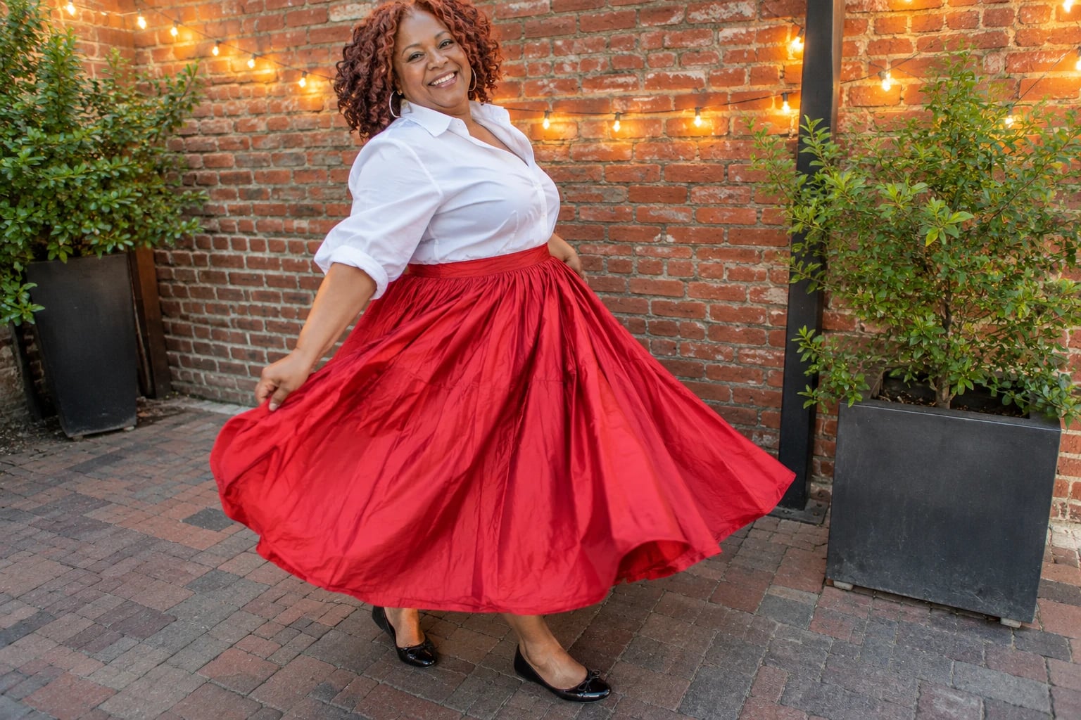 A plus-size woman twirls a full red taffeta skirt over black flats near a brick wall strung with lights.