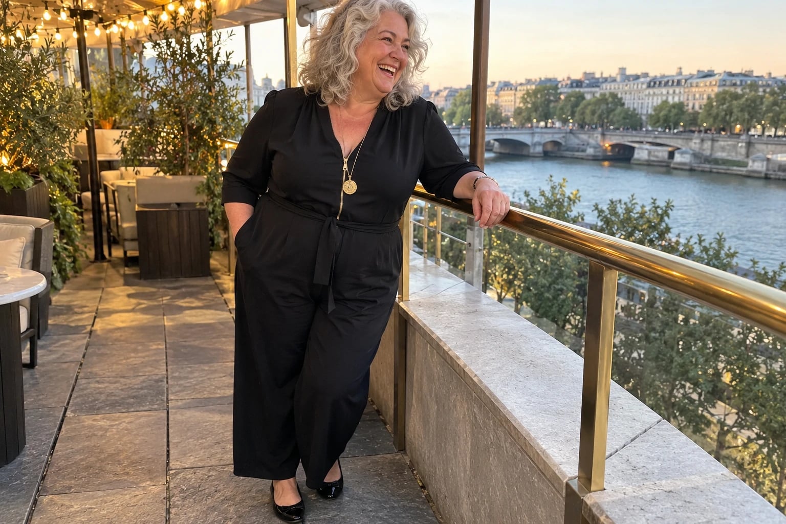 A plus-size woman in her fifties laughs on a terrace railing, wearing a black wide-leg jumpsuit and gold pendant necklace.