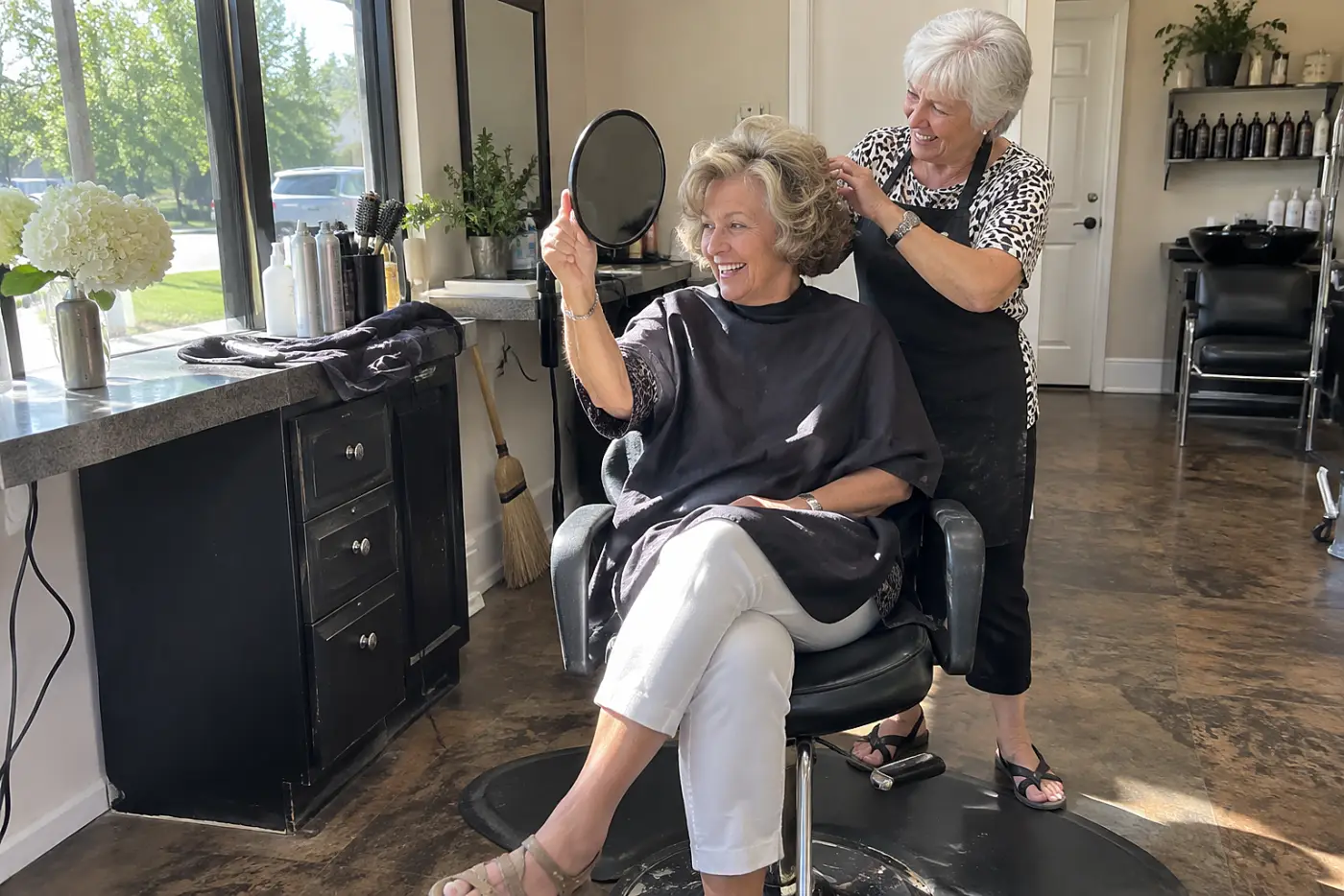 A woman in her 60s holds a hand mirror behind her head while seated in a salon chair.