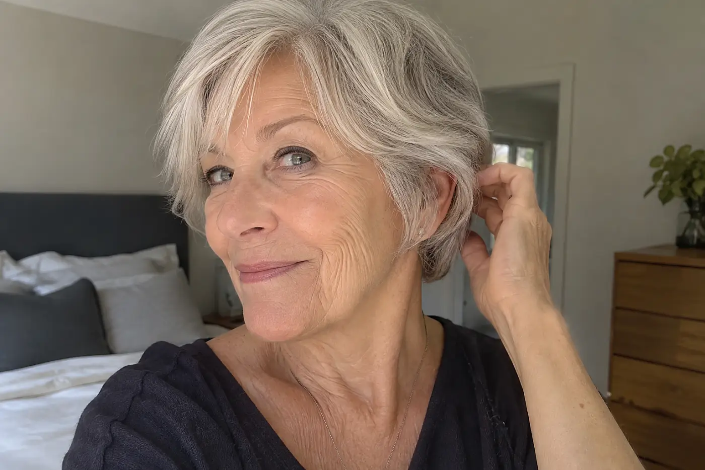 Woman brushing the rounded edge of her silver pixie away from her cheek in a bedroom.