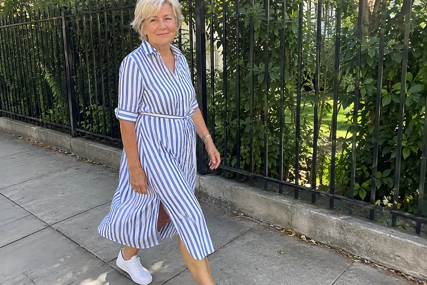 Woman in her sixties in a blue-and-white striped shirtdress and white sneakers climbing metal bleachers at a youth baseball field.