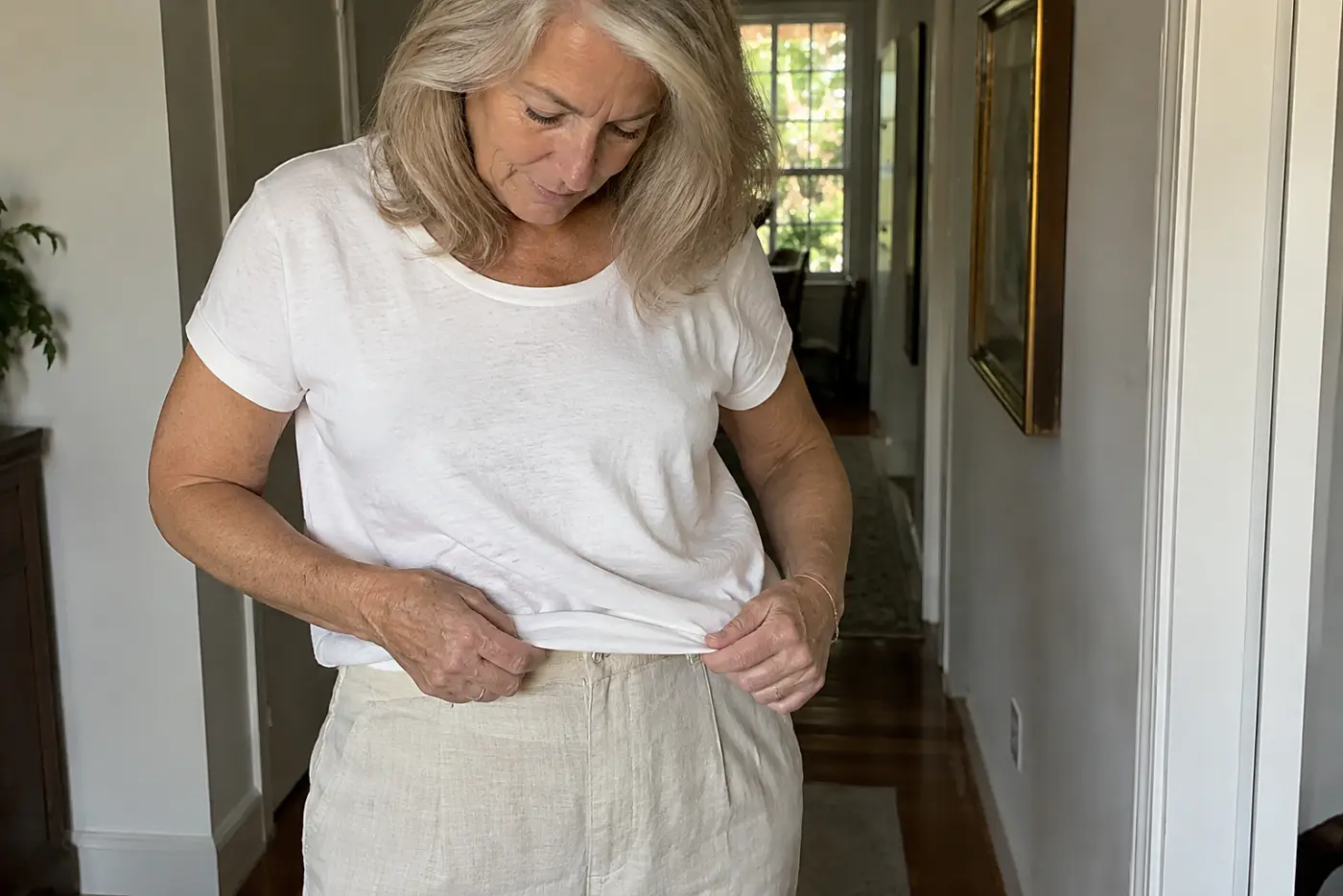 Woman in her sixties in linen trousers carrying a basket of herbs across a sunny backyard.