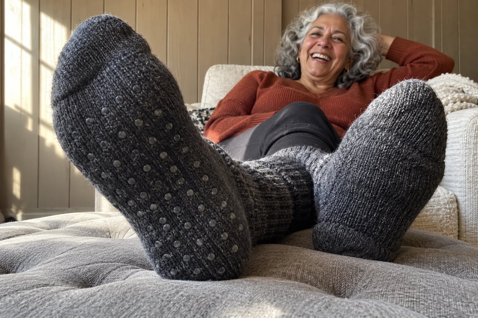 A pair of thick charcoal gray wool socks with small rubber dots on the soles, one foot turned to show the grip, is worn by a woman in her sixties resting her feet on an ottoman in her den.