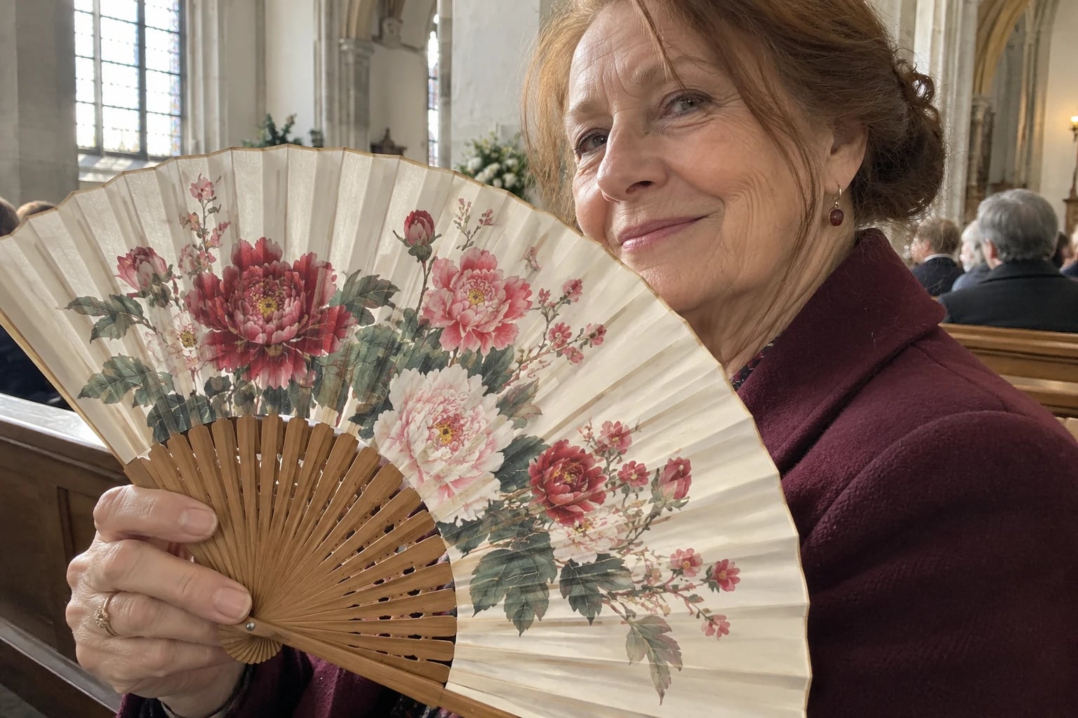 A painted paper fan with a floral pattern and no lettering is opened wide in the hand of a woman in her fifties sitting in a wooden church pew on a winter afternoon.