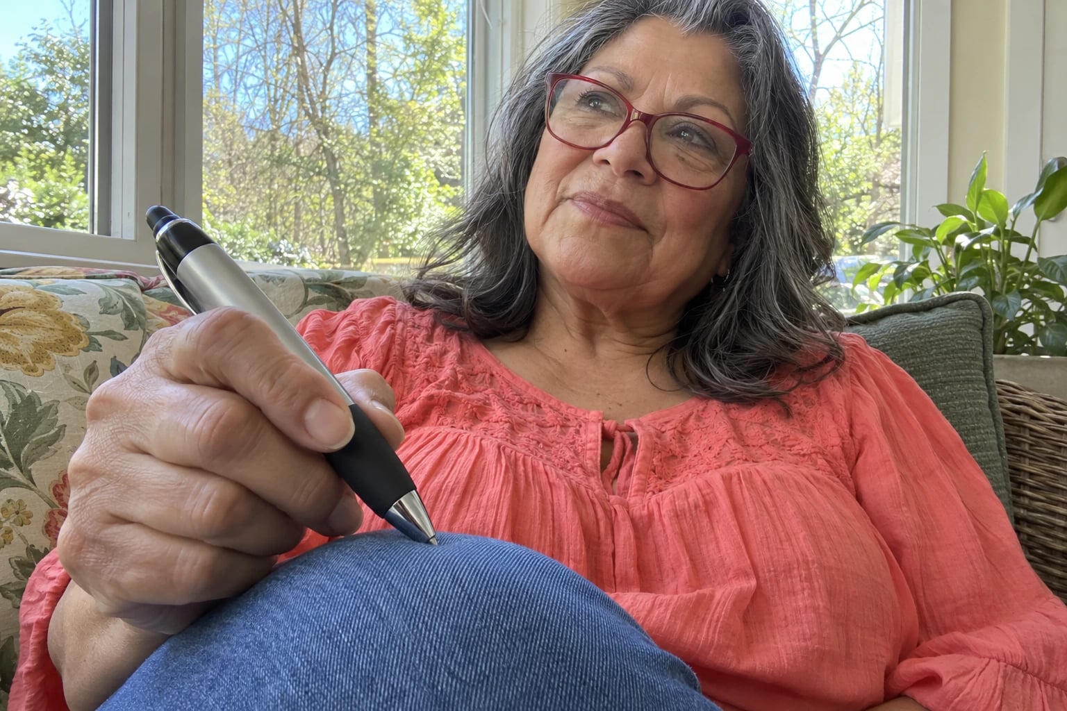 A gray-haired woman in red glasses and a coral blouse holds a chunky silver pen on her knee.