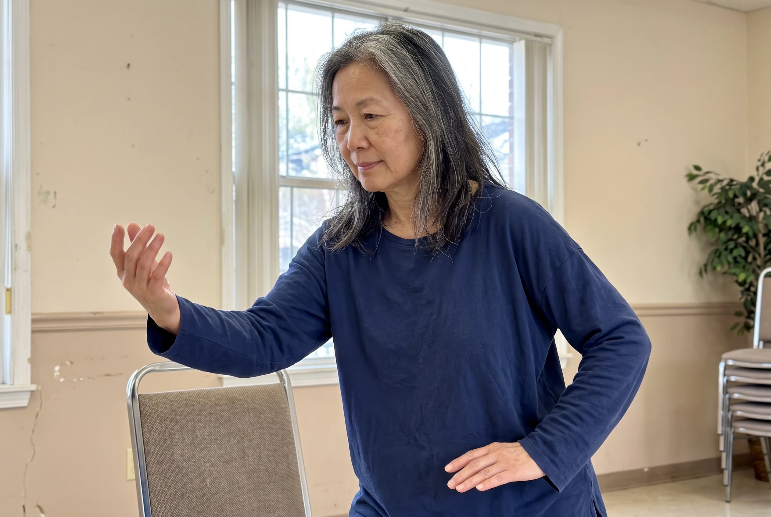 Woman practicing Parting the Wild Horse’s Mane beside a chair in a community room.