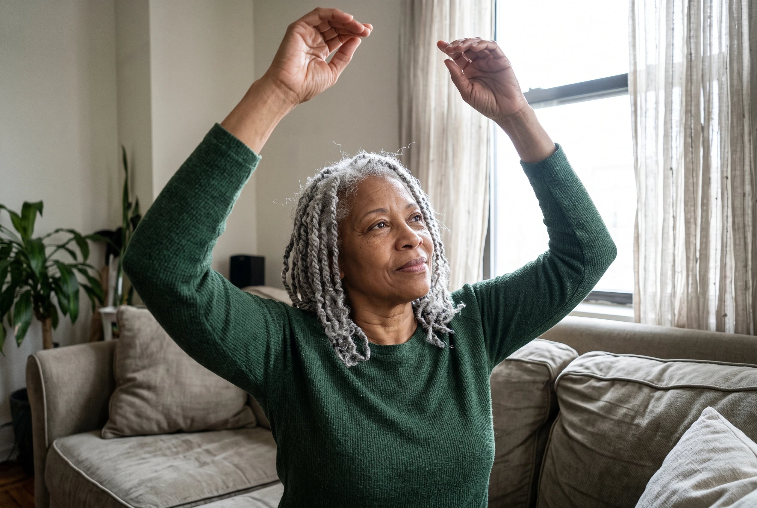 Woman raising both hands overhead beside a sofa in her living room.