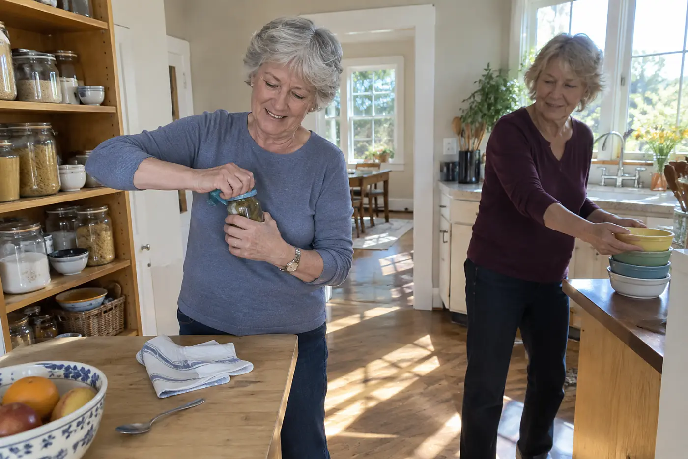 A woman in her sixties opens a jar with a blue rubber grip in a kitchen.