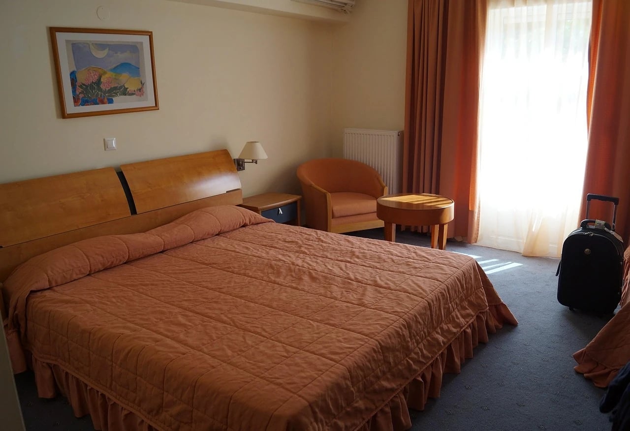 Hotel room with a double bed, orange bedspread, armchair, round table and a black suitcase by the window
