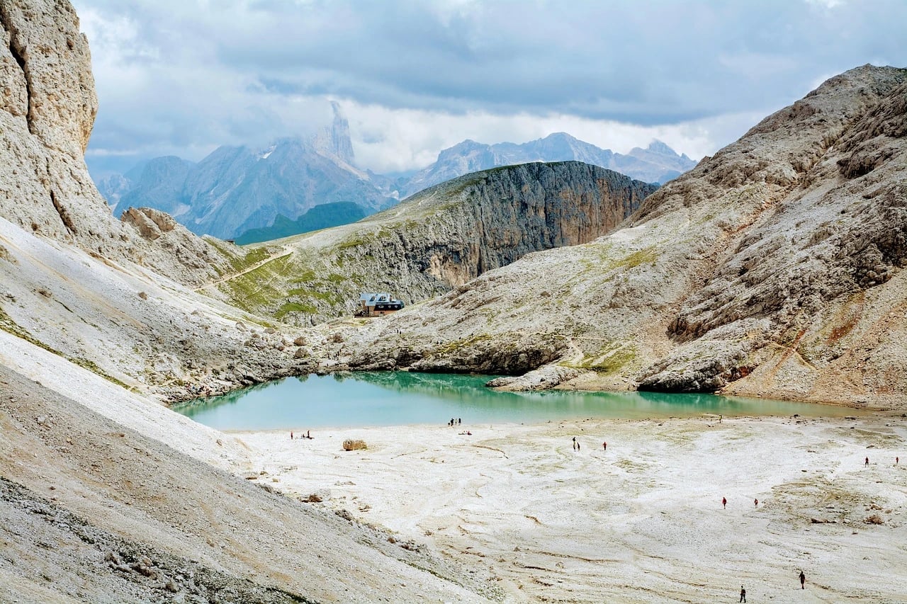 Turquoise alpine lake and mountain hut in the Dolomites with tiny hikers walking across pale rocky ground