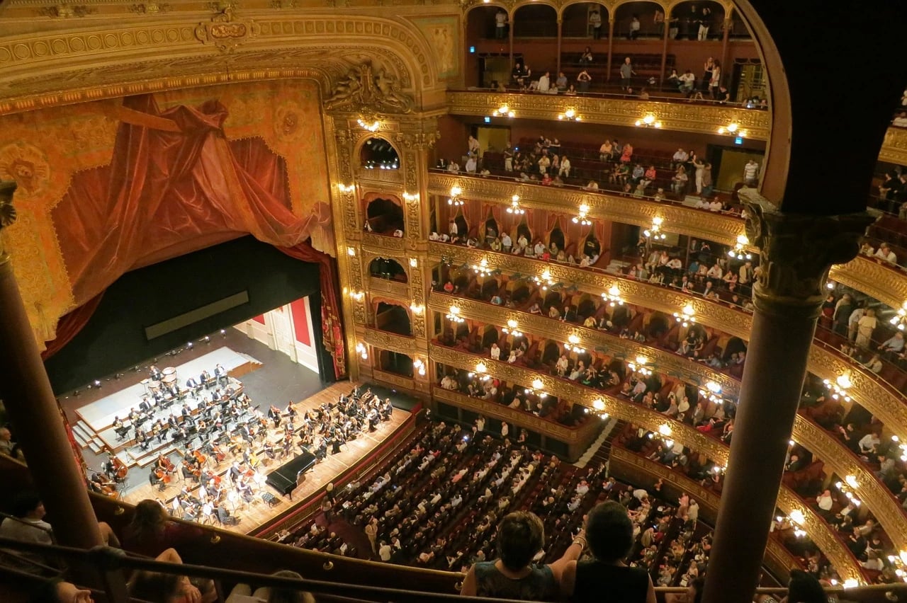 Ornate multi-tiered opera house interior seen from the upper balcony, with a full audience and orchestra on stage