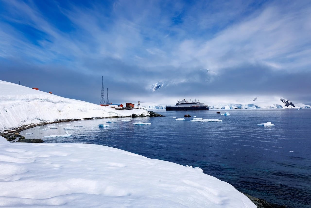 Dark expedition cruise ship anchored in a calm icy bay with snowy slopes, research buildings and small boat
