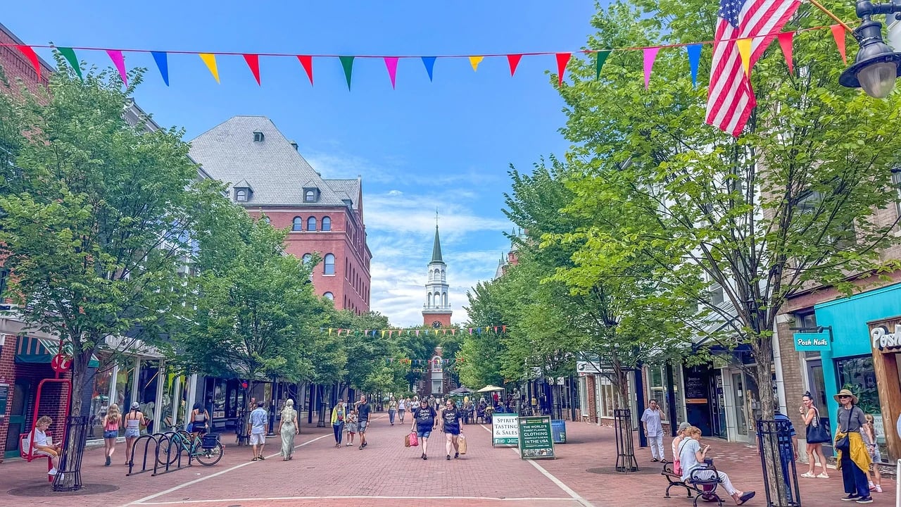 Pedestrians walking along the brick Church Street Marketplace in Burlington, Vermont, with shops, trees, bunting and a church steeple ahead