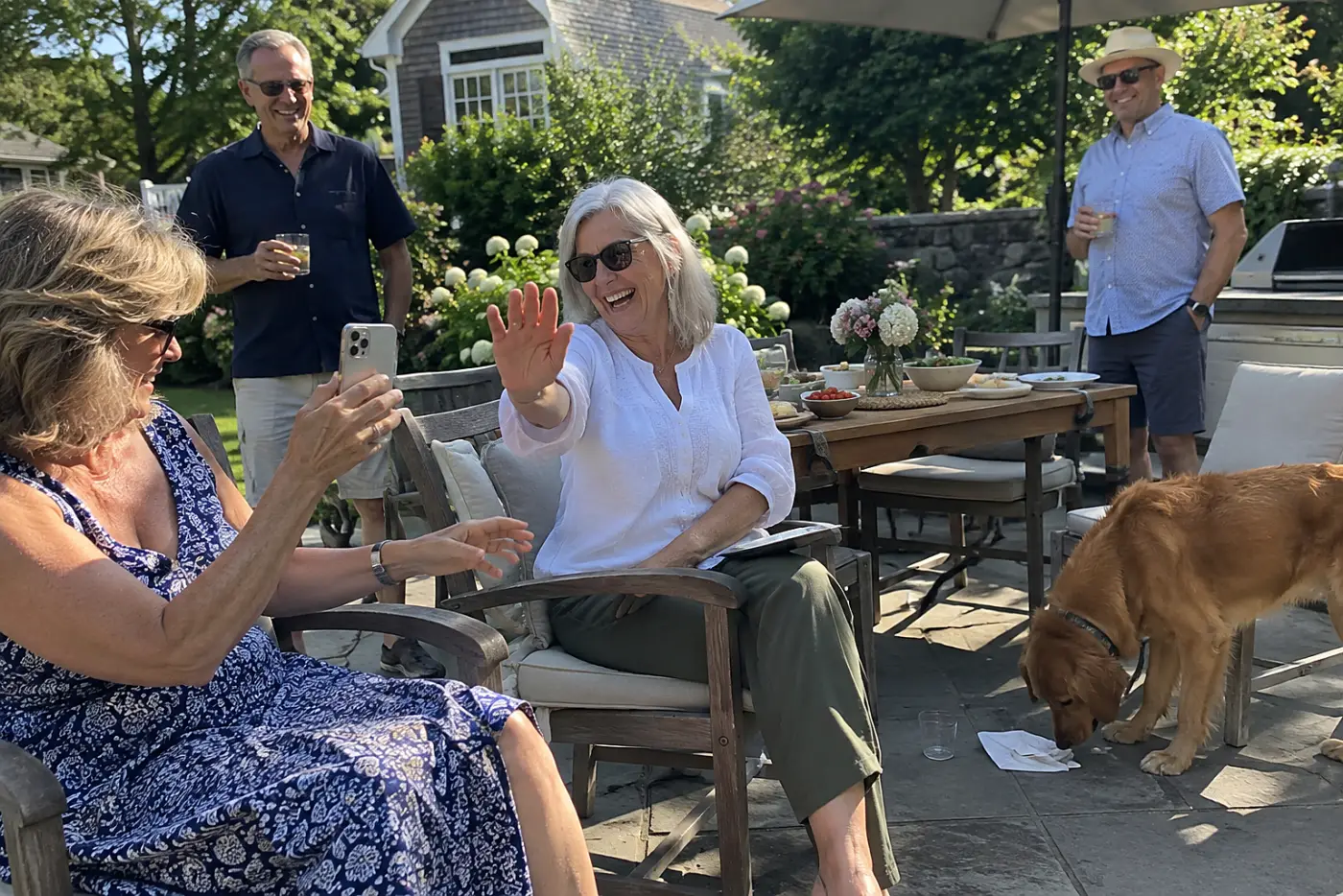 A woman in her sixties lowers her hand between a friend's phone and her face at an outdoor garden party.
