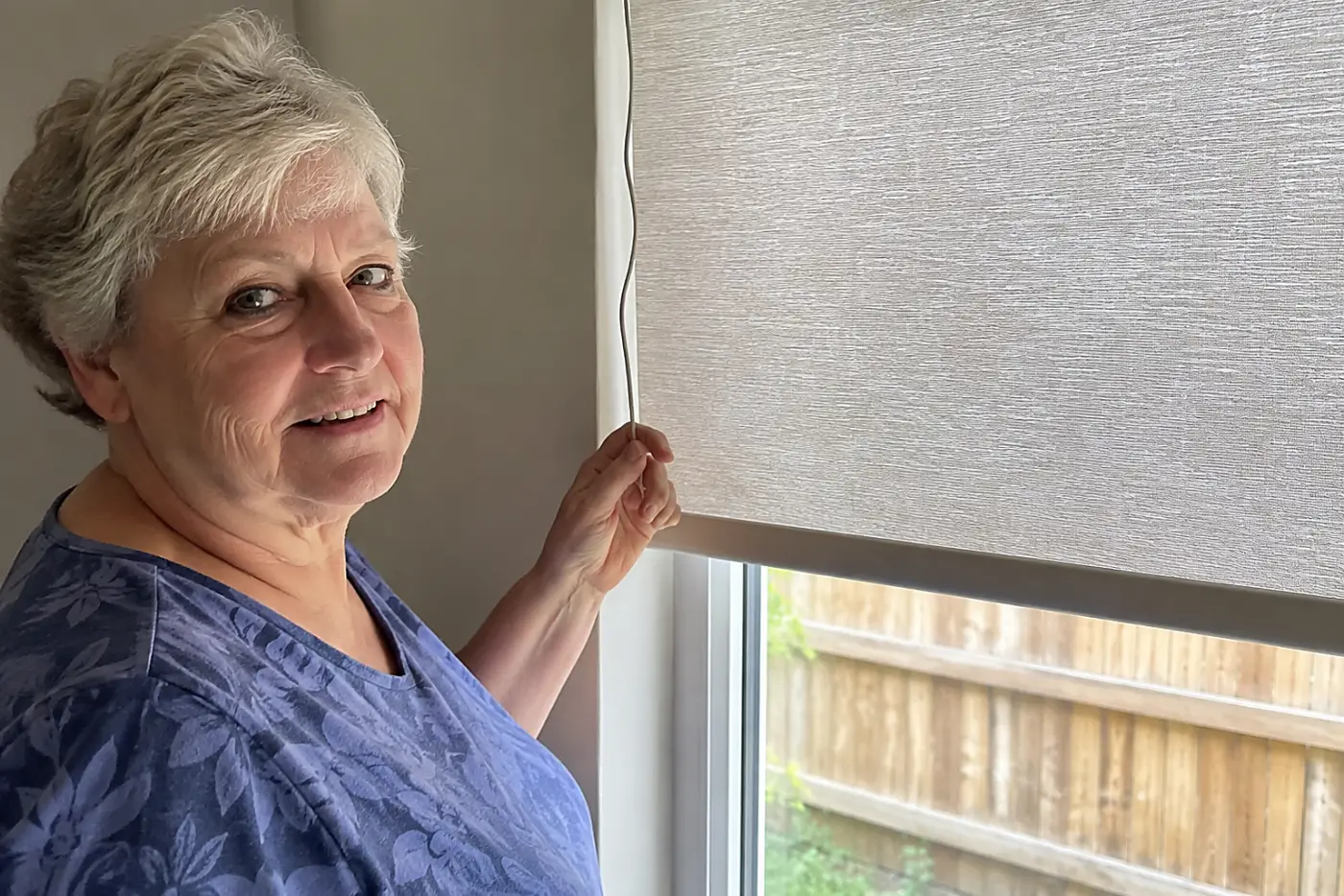 A window shade cord fills the center of a real living-room window as a woman over 70 stands beside the lowered shade, with no writing visible.
