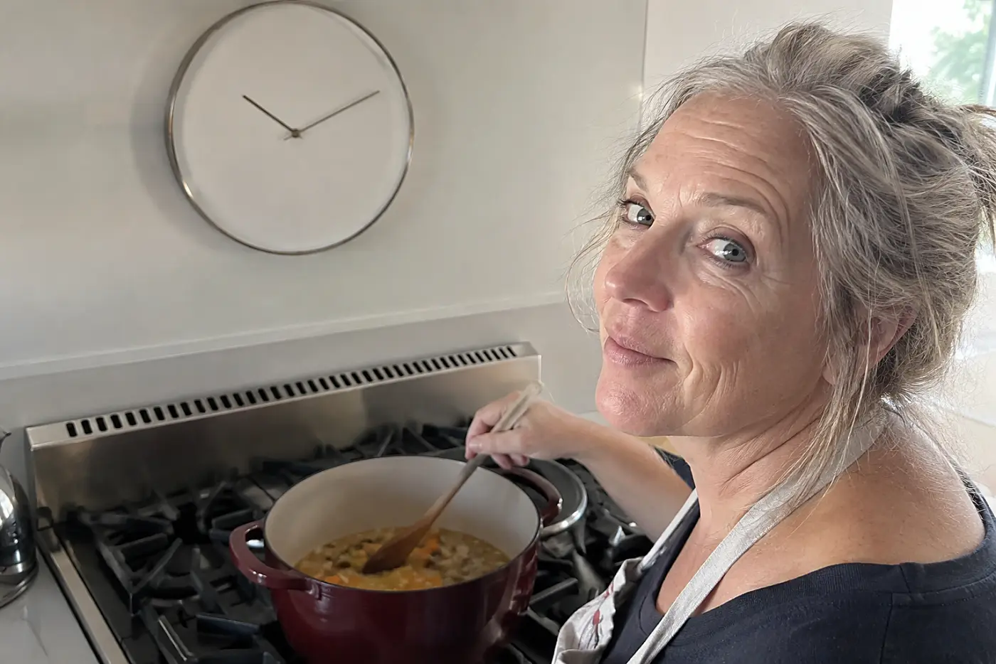A round clock hangs in the middle of a kitchen wall above the stove as a woman over 50 checks it while stirring a pot, with no visible writing.