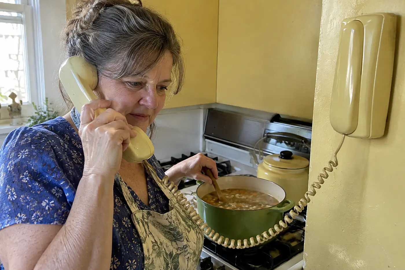 A wall phone with a long cord hangs in the middle of a 1970s kitchen wall as a woman over 50 holds its handset by the stove, with no visible writing.