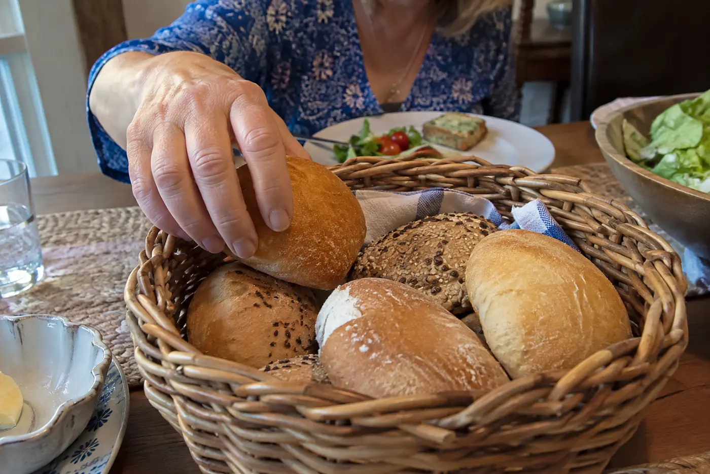 A wicker bread basket sits in the middle of a family supper table as a woman over 50 reaches for a roll, with no visible writing.