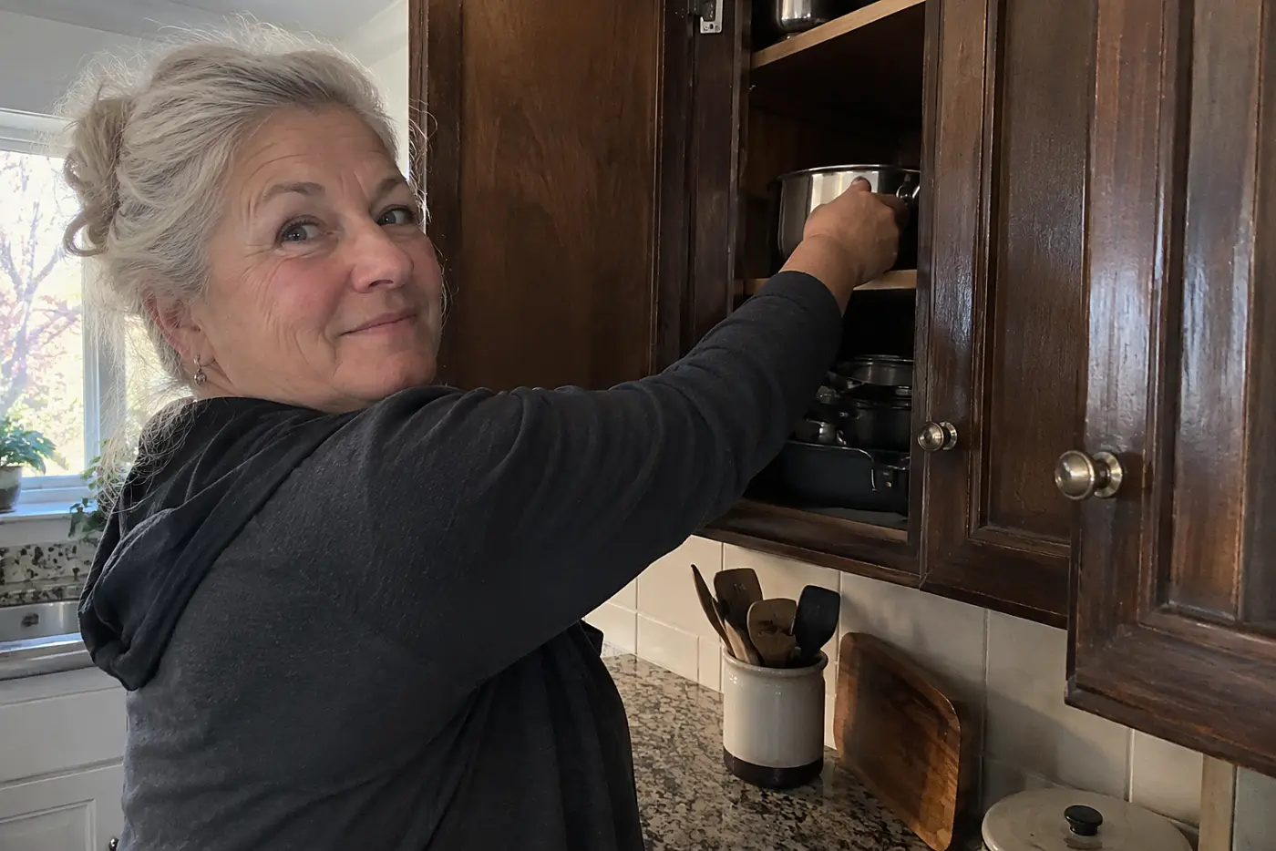 Dark wood cabinet doors fill the middle of a kitchen as a woman in her sixties reaches for a saucepan, with no visible writing.