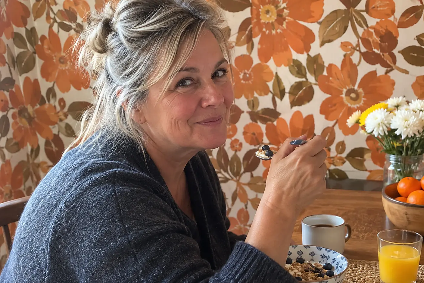 Orange-and-brown floral wallpaper fills the middle of a kitchen wall behind a woman over 50 seated at breakfast, with no visible writing.