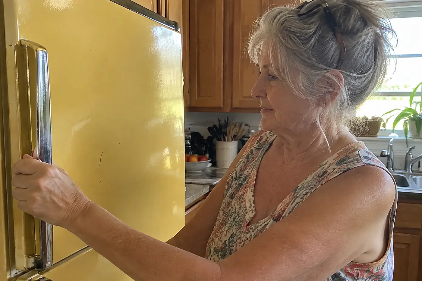 A harvest-gold refrigerator fills the middle of a lived-in kitchen as a woman in her sixties reaches for its handle, with no visible writing.