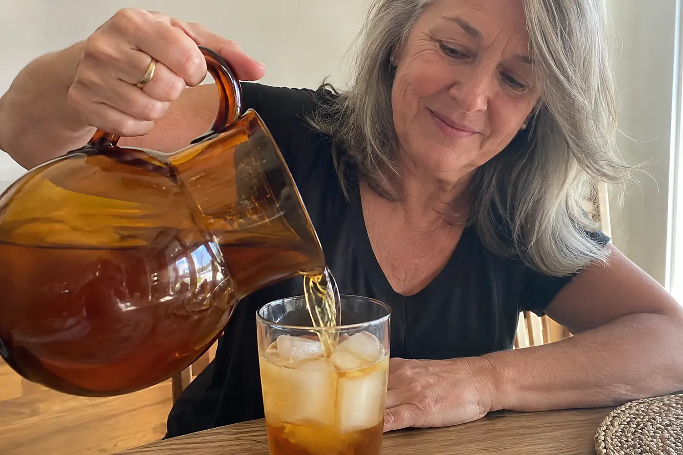 A brown glass pitcher sits in the middle of a kitchen table as a woman over 50 pours iced tea, with no visible writing.