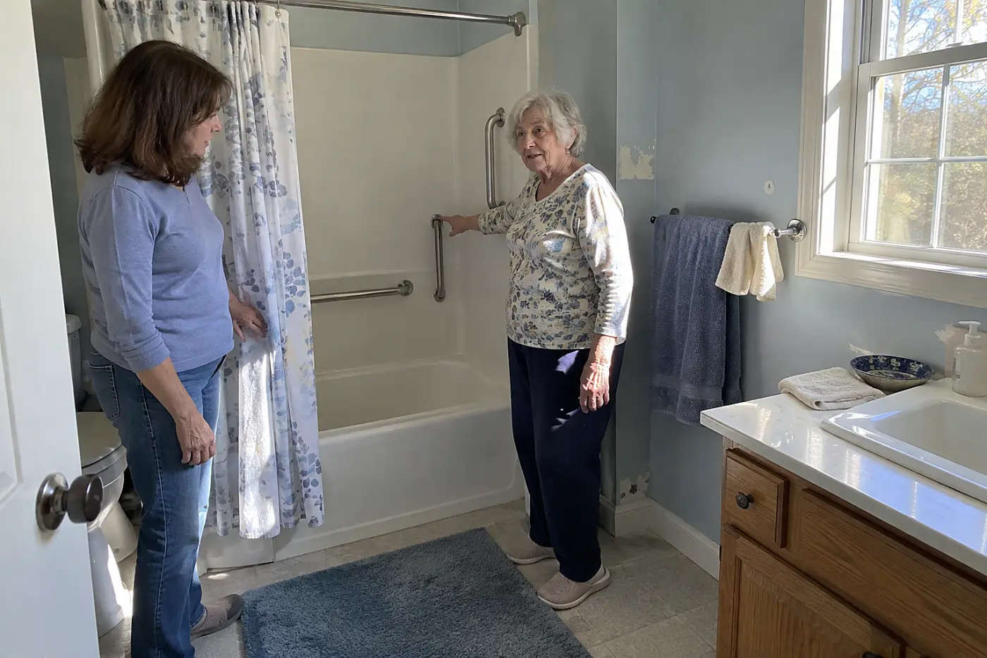 Older woman pointing to a wall beside her bathtub where she reaches for support.