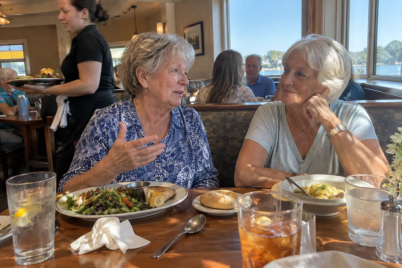 Older woman leans toward a friend across a busy restaurant table.