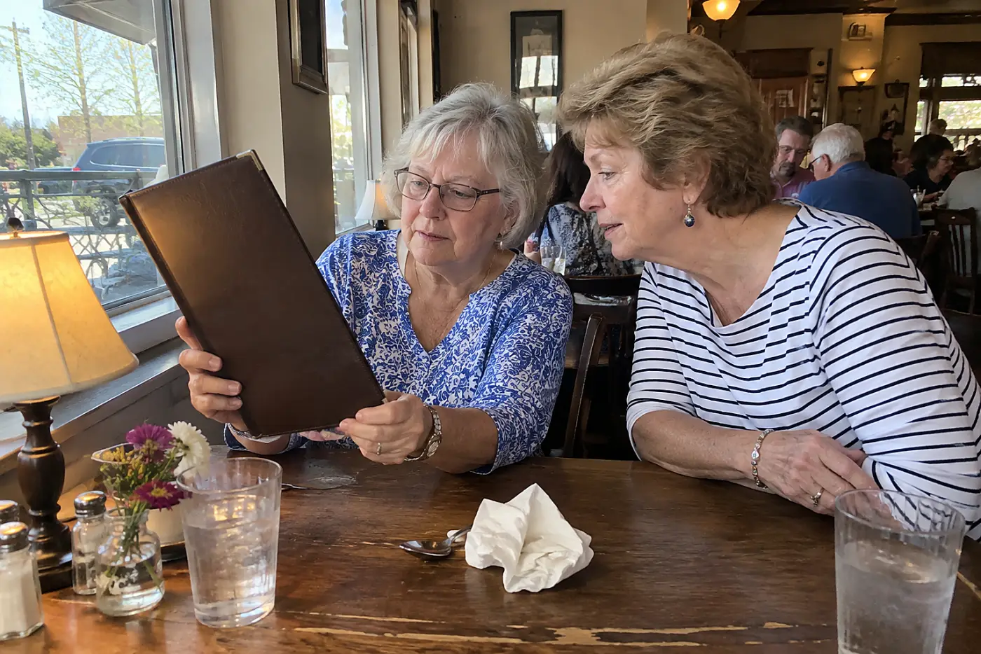 Older woman holds a restaurant menu toward a lamp at a corner table.