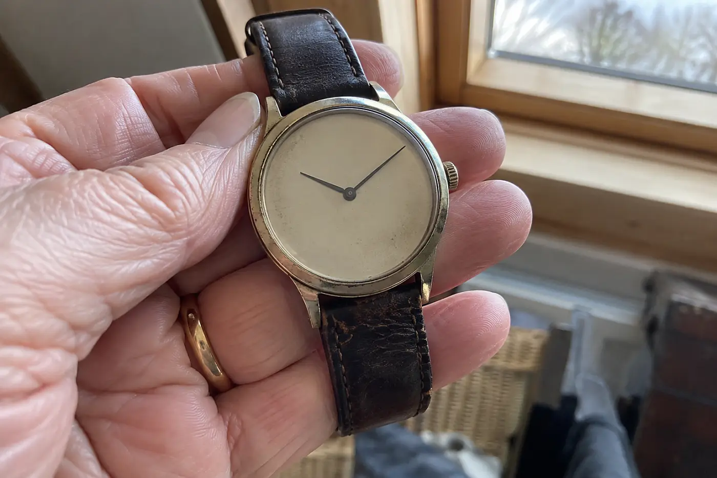 A stopped mechanical wristwatch rests in the middle of a woman's palm beside an attic window, with no visible writing.