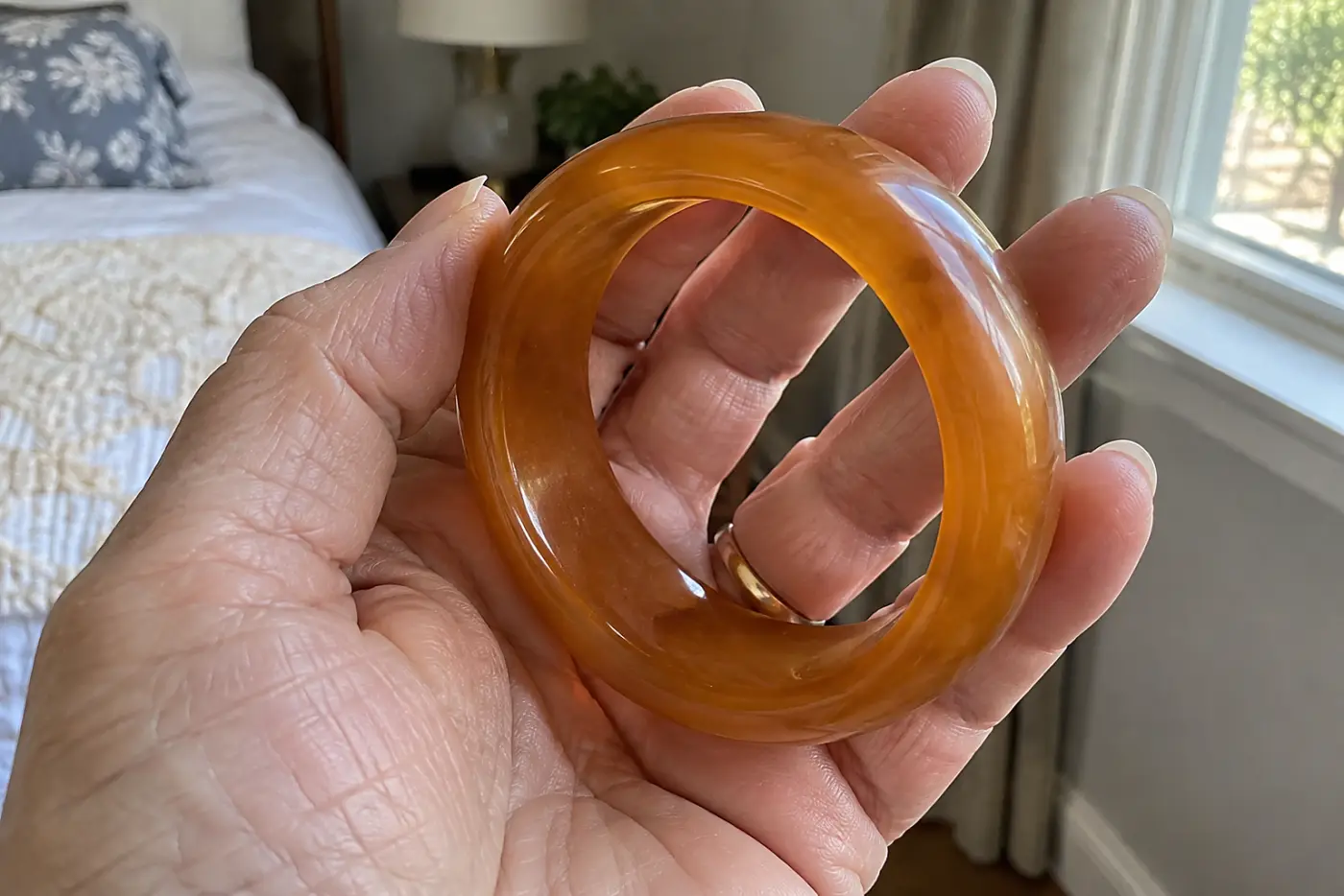 A thick amber Bakelite bangle sits in the middle of a woman's hand by a bedroom window, with no visible writing.