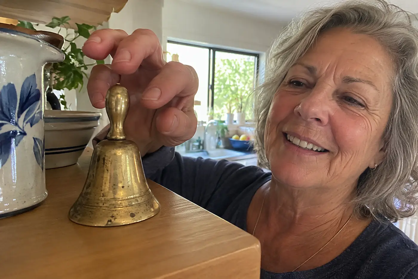 A small brass school bell sits in the middle of a kitchen shelf as a woman in her sixties reaches for it, with no visible writing.