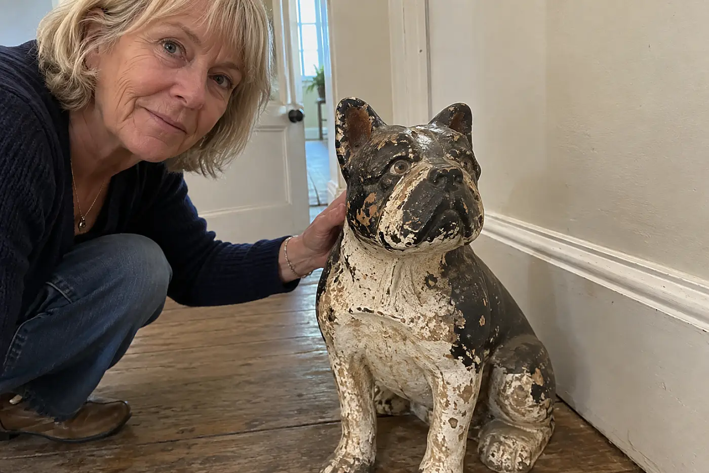 A chipped cast-iron dog doorstop sits in the middle of a worn hallway floor near a woman in her sixties, with no visible writing.