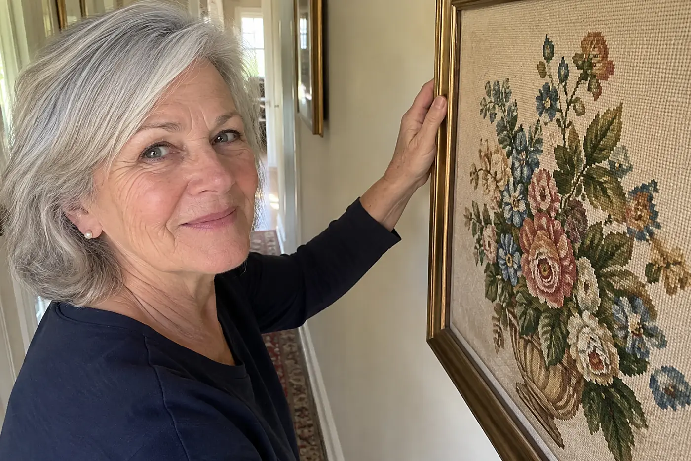A framed needlepoint hangs in the middle of a lived-in hallway wall beside a woman in her sixties, with no visible writing.