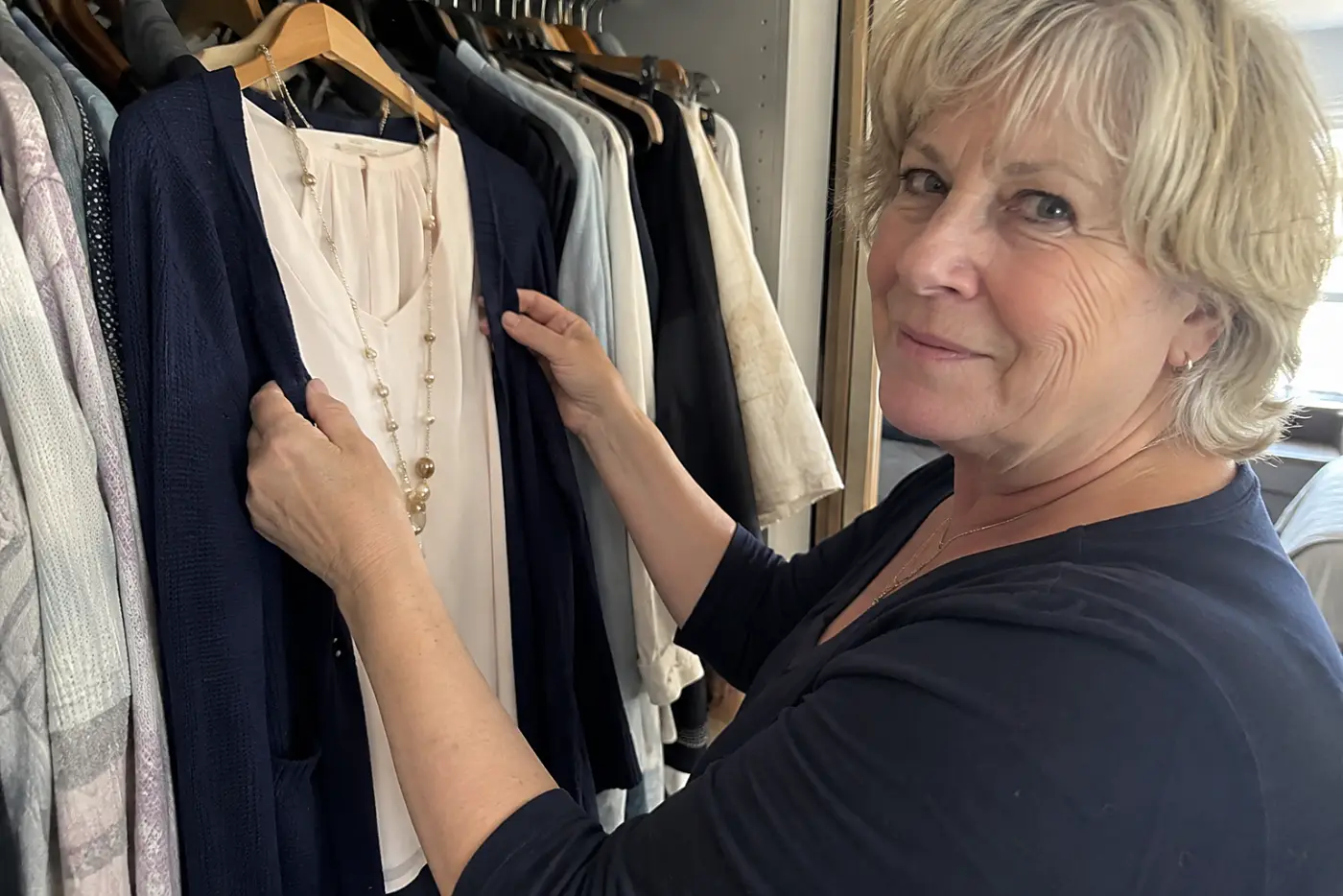 A blouse, cardigan, and necklace hang together on one hanger in the center of an open closet as a woman in her sixties reaches to separate them, with no writing visible.