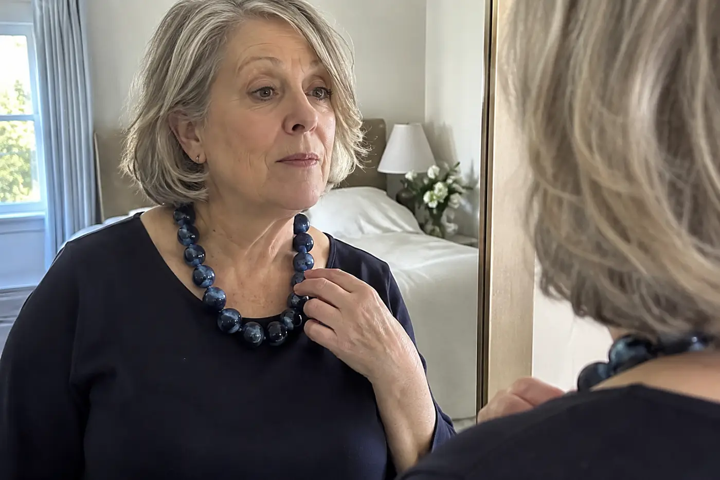 A large glossy-bead necklace sits at the center of a woman in her sixties' neckline as she checks it in a bedroom mirror, with no writing visible.