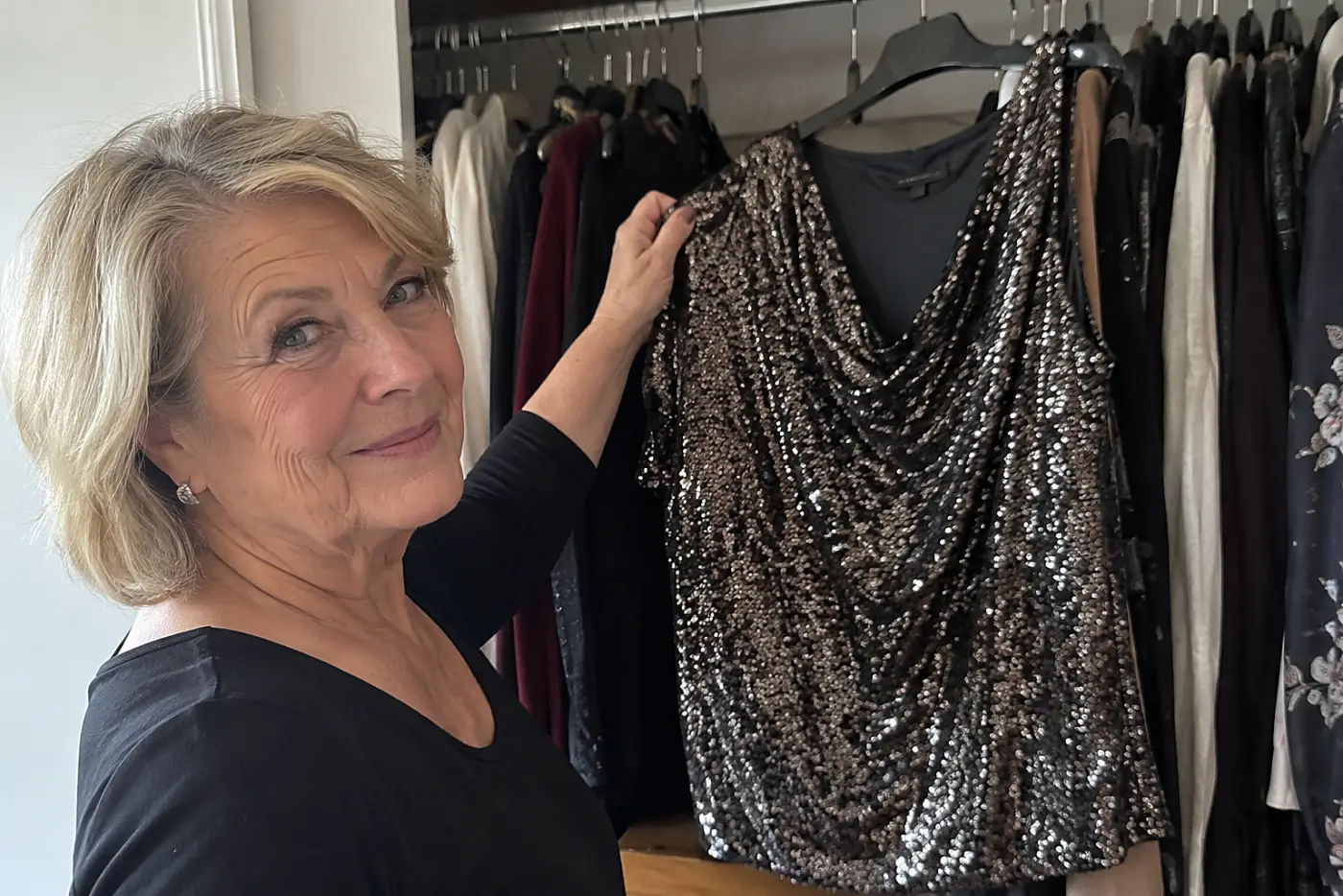 A sequined evening top hangs in the center of an open bedroom closet as a woman in her sixties checks its shoulder, with no writing visible.
