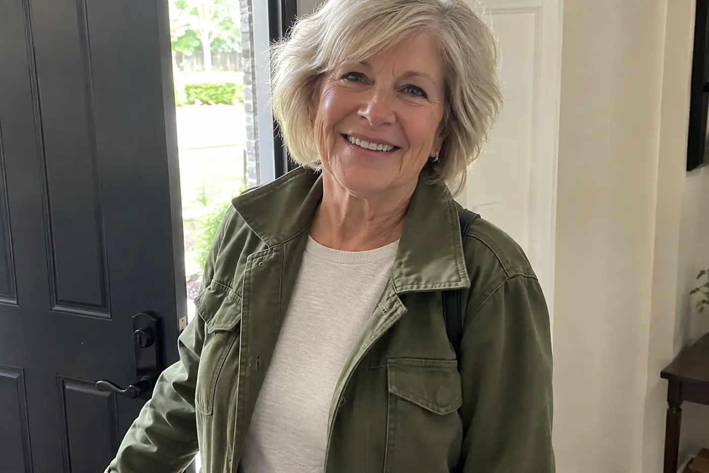An olive green jacket sits open in the middle of the frame on a woman in her sixties standing in her entryway.
