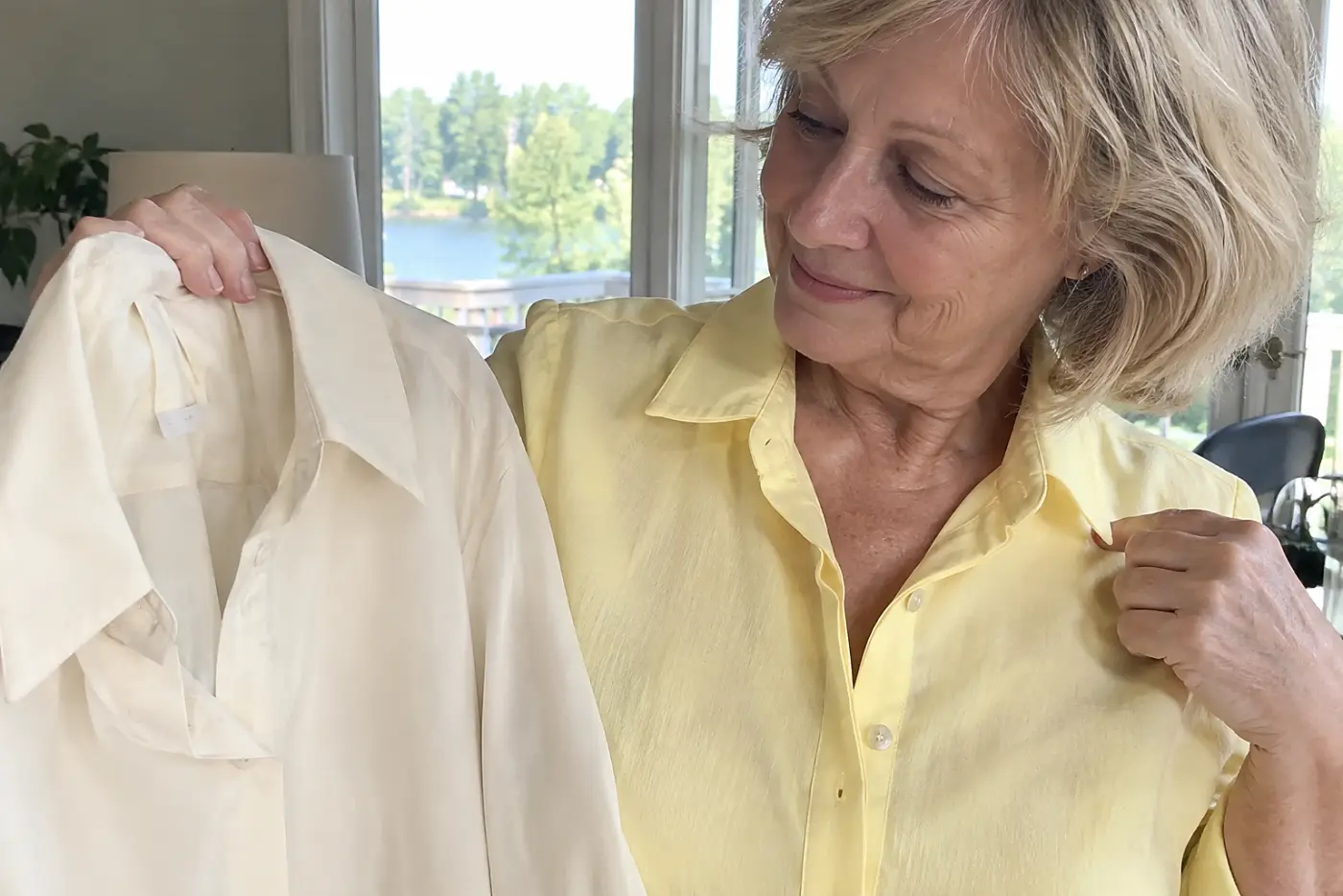 A butter yellow blouse fills the middle of the frame on a woman in her sixties holding a plain cream blouse beside a window.