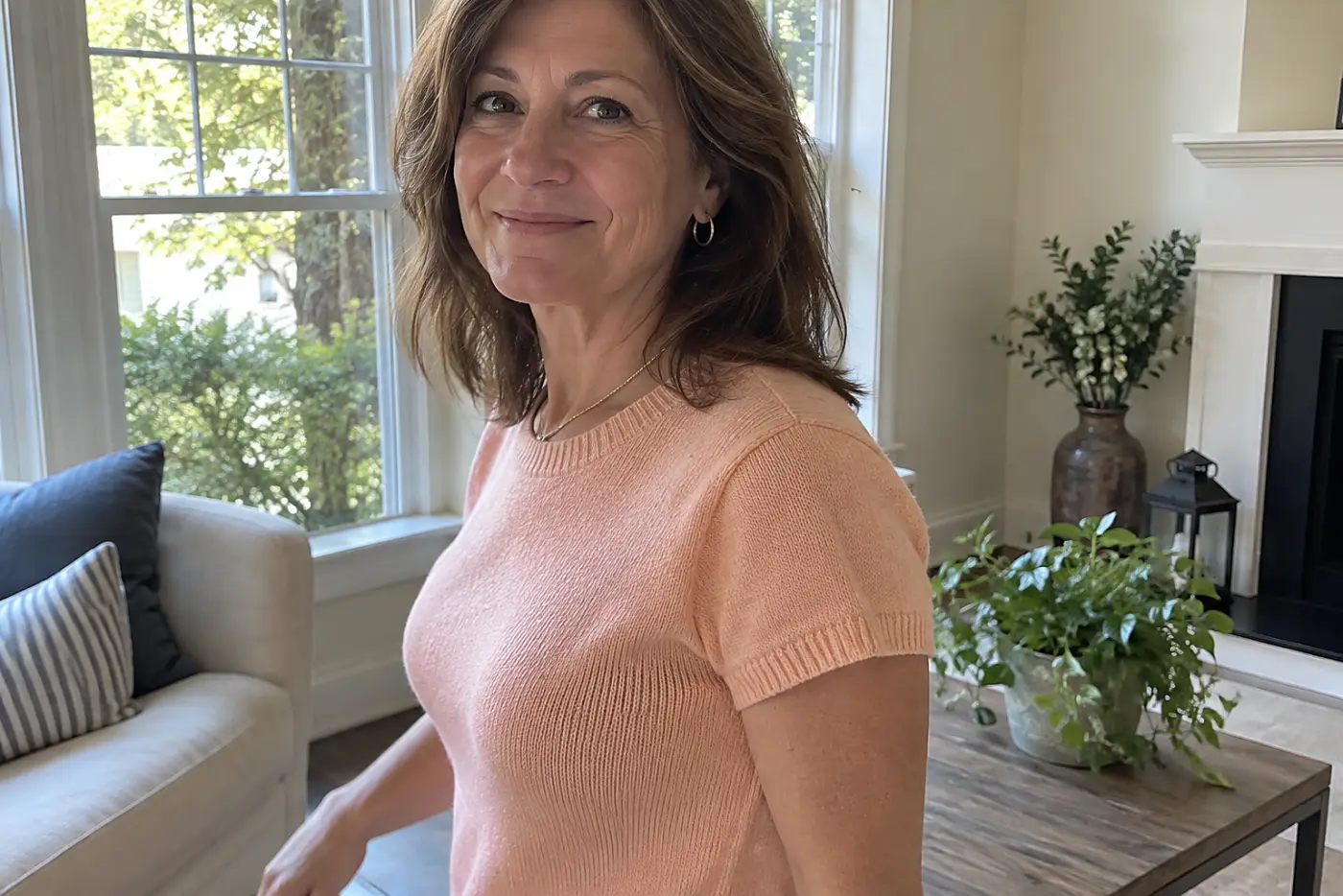 A peach knit tee fills the middle of the frame on a woman in her fifties standing near her living-room window.