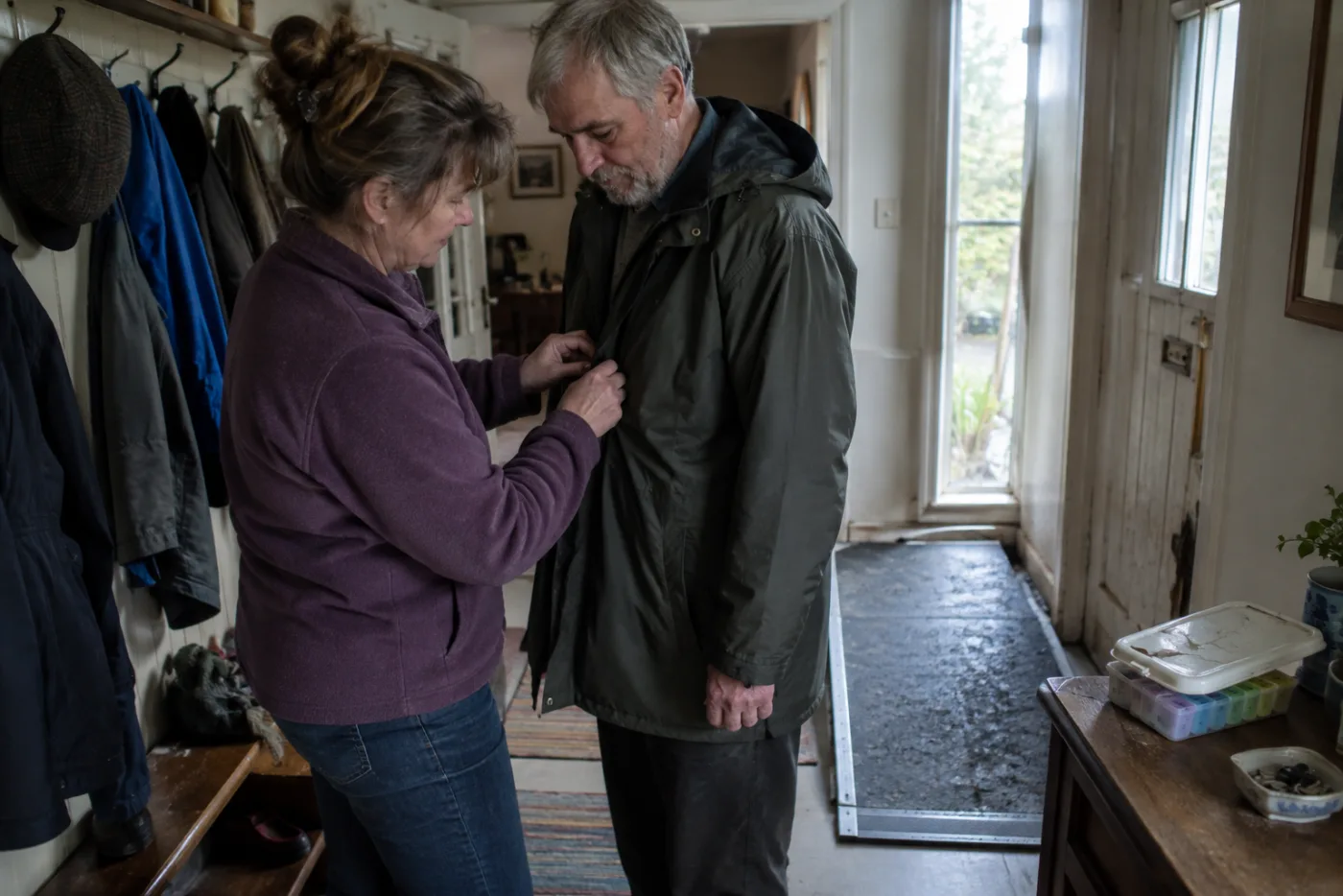 Woman buttoning her brother's raincoat in a hallway with a wheelchair ramp mat