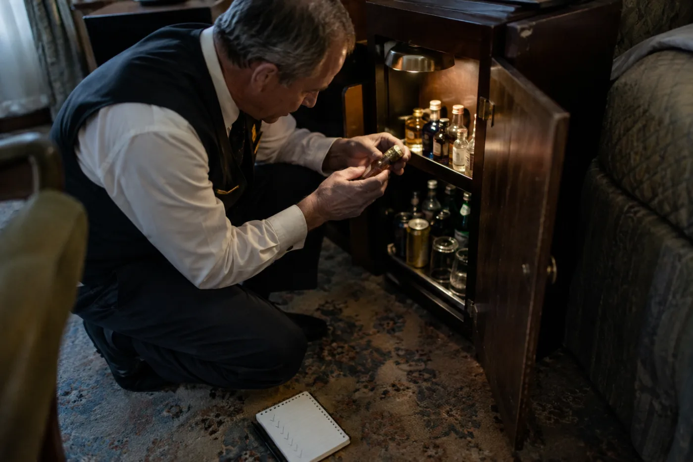 Steward inspects a minibar bottle's foil seal beside a small cabin cabinet.