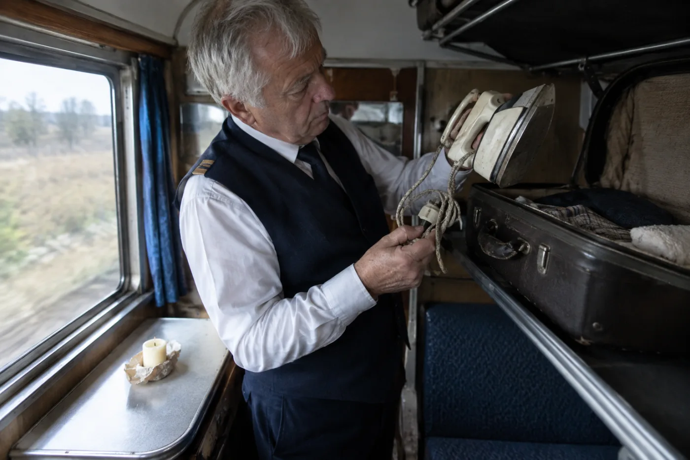 Steward lifts a travel iron from a suitcase, candle beside it on desk.