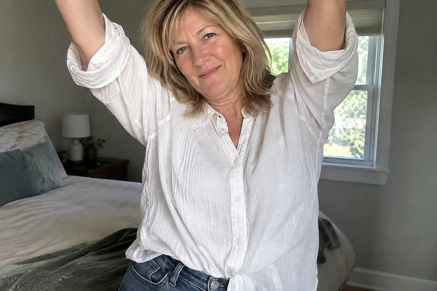 A loosely tucked shirt is centered on a woman over 50 raising both arms in her bedroom.