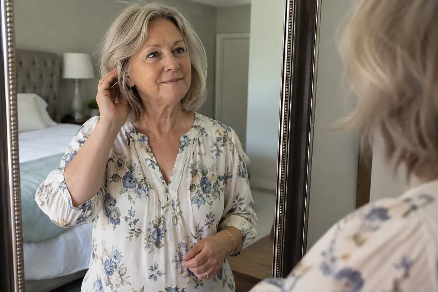 A shirt with shoulder seams in place is centered on a woman in her sixties looking at her reflection in a bedroom mirror.