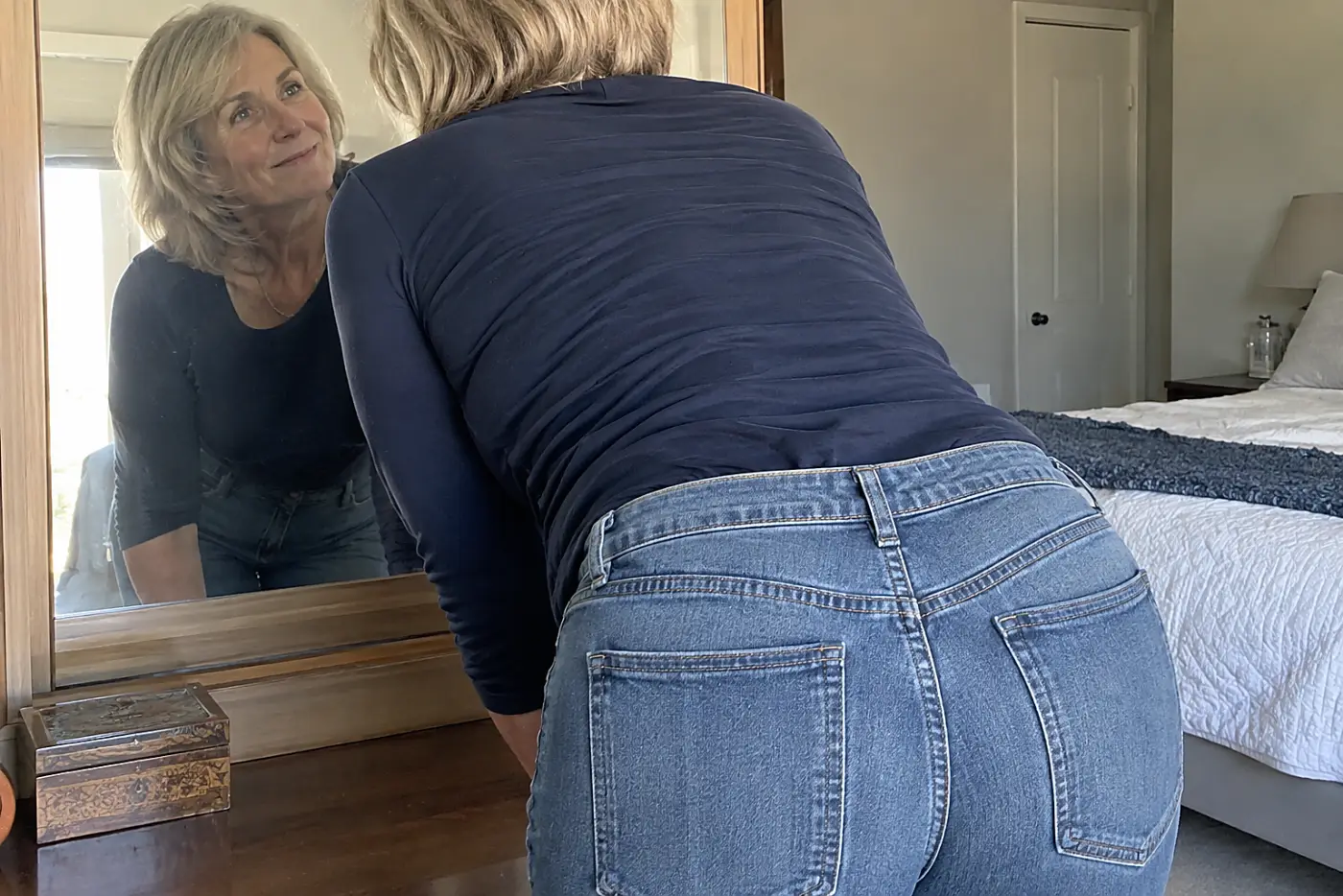 Good jeans with a tailored waist are centered on a woman in her sixties bending toward a bedroom mirror, with the back waistband visible.